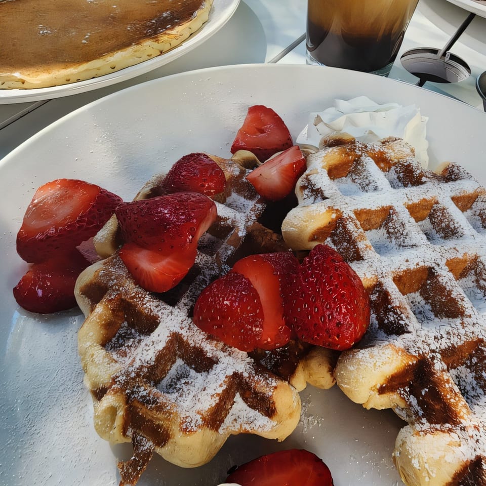 Golden waffles with powdered sugar and fresh strawberries, alongside a pancake and iced coffee.